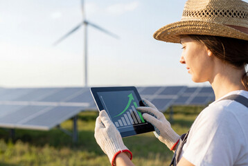 Fototapeta premium Female technician analyzing growth chart on tablet at solar farm with wind turbine at sunset, clean energy monitoring and sustainable power management for renewable industry operations