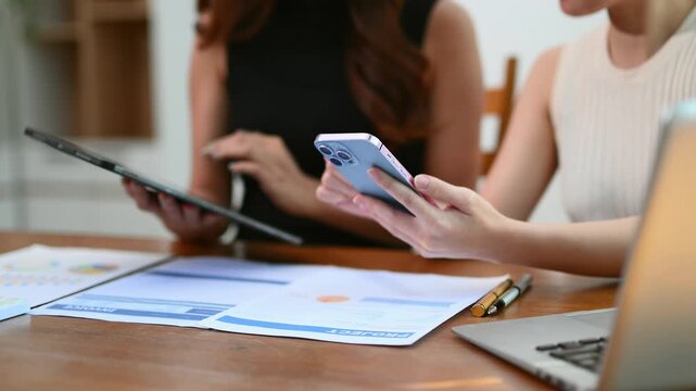 Two women collaborating in a modern office, discussing data, digital planning, and business strategy using tablet, smartphone, and documents.