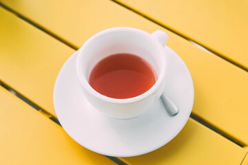 A white cup of hot herbal or fruit tea sits on a bright yellow table, creating a clean, minimalistic, and refreshing composition with vibrant contrasting colors.