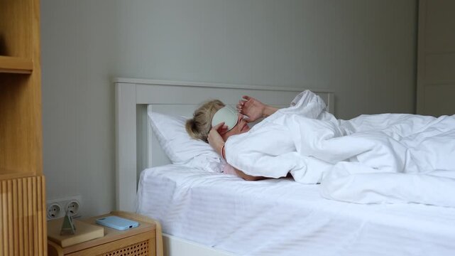 Sleepy positive middle aged woman waking up lying in comfy bed, peeks out from mask and continues to sleep wrapping in blanket. Female being lazy to get up, sleeping in early morning in cozy bedroom.