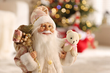 Santa Claus figure with sack of gifts and toy bear against blurred lights indoors, closeup. Merry Christmas and Happy New Year