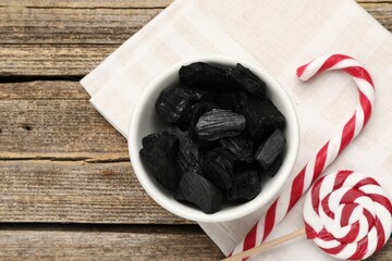 Pieces of black coal in bowl, Christmas candy cane and lollipop on wooden table, flat lay. Space for text
