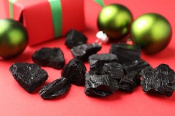 Pieces of black coal, gift box and Christmas baubles on red background, closeup