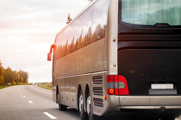 Large coach bus travels on asphalt road during sunset, warm sunlight reflects on vehicle side,...