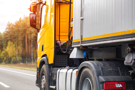 Yellow semi truck trailer movement on highway. Side view transport vehicle travel asphalt road with forest background. Early morning or late afternoon sun glow.