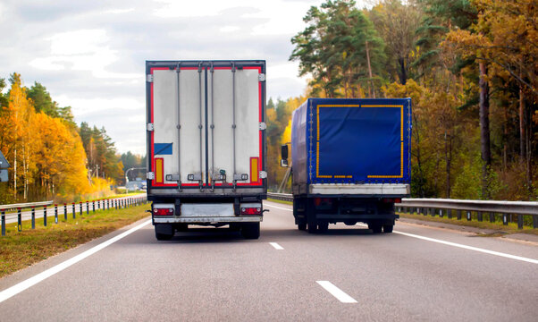 Rear view of two trucks driving on asphalt road during autumn day. White semi-trailer and blue curtain-side truck transport cargo on highway passing colorful forest under cloudy sky.