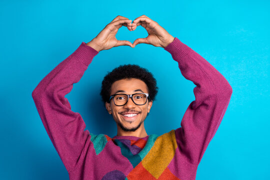 Trendy man in colorful sweater gesturing heart sign with hands against bright blue background, showing happiness.