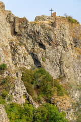 A rock covered with faded grass with a wooden cross. An iron staircase leads to the top. The remains of a fortress are visible. Green bushes in the foreground. Azeula fortress. Kojori