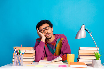 Funny male student resting during study surrounded by books and sitting at desk with vibrant blue background