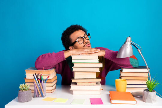 Young male student leaning on stacked books with a thoughtful gaze at a desk surrounded by bright stationery on blue background