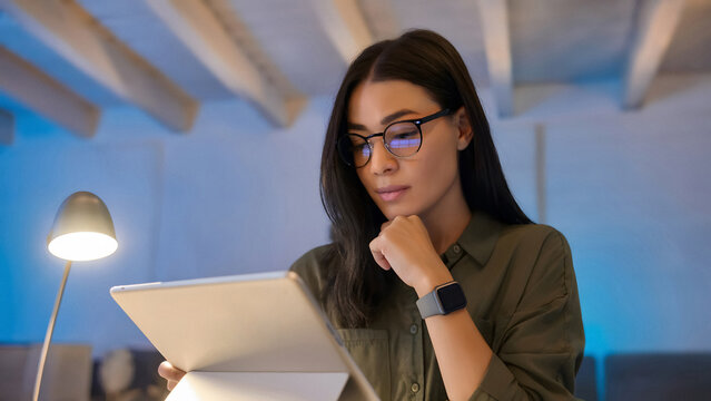 focused woman working on digital tablet at home office desk with warm lamp light in modern workspace
