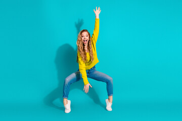 Young happy girl in yellow sweater jeans and white sneakers jumping with arms raised against blue...