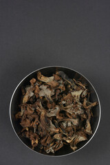 A stainless steel bowl full of dried, dark Black Trumpet mushrooms is centered on a solid dark gray background in an overhead shot