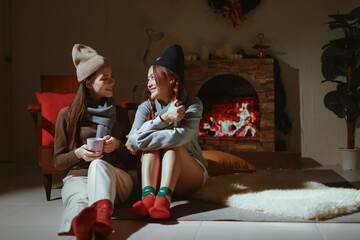 Teen girls having fun together on holiday, sitting in front of the fireplace. Asian lesbian couple celebrating Christmas, drinking warm drinks to keep warm.