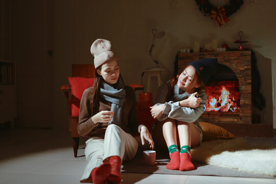Teen girls having fun together on holiday, sitting in front of the fireplace. Asian lesbian couple celebrating Christmas, drinking warm drinks to keep warm.