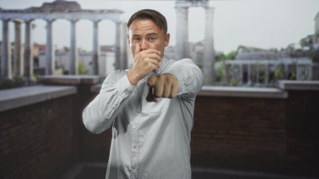 Man points finger at camera, laughs and alternately covers mouth while wearing a light patterned shirt on a building rooftop terrace with brick parapet and classical columns; playful confidence.