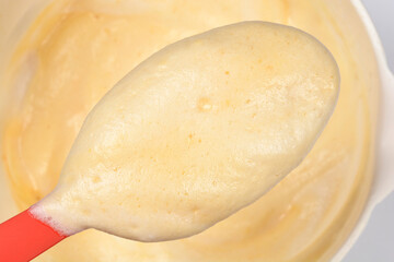 A close-up shows a red spatula lifting a generous spoonful of light yellow, airy sponge cake batter, highlighting its smooth, frothy texture