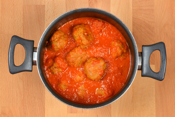 An overhead shot captures a silver cooking pot filled with meatballs submerged in a rich, vibrant red tomato sauce, resting on a light wooden surface