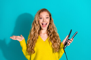 Young female model with long hair smiles holding a hair straightener in a bright blue studio