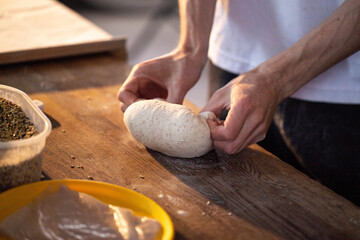 Close up of hands of baker or home cook shaping piece of artisanal bread dough on rustic wooden countertop with natural light highlighting texture of flour and grains