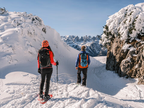 Two hikers with backpacks and snowshoes stand on a snowy ridge, looking at distant mountains, Brenta Dolomites. Paganella, Roda refuge,Trentino, Italy