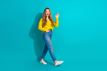 Young woman waving in a yellow sweater jeans and white sneakers against a blue background