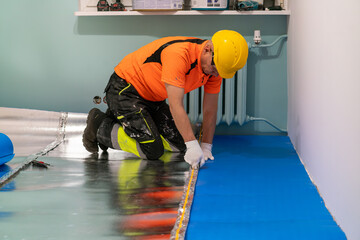 A finishing worker applies an insulating underlayment with an aluminum layer under wooden floor panels. Sound and thermal insulation. Apartment renovation.