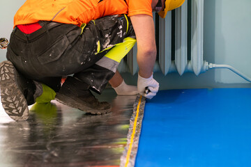 A finishing worker applies an insulating underlayment with an aluminum layer under wooden floor panels. Sound and thermal insulation. Apartment renovation.