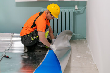 A contractor applies an insulating underlayment with an aluminum layer under wooden floor panels. Sound and thermal insulation. Apartment renovation.