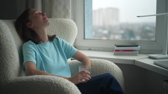 A woman sits comfortably in a chair by the window, practicing relaxation techniques to relieve stress. The afternoon light fills the room, creating a calm atmosphere.