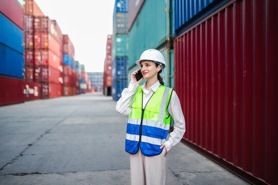 A woman in protective gear talks on the phone, addressing important logistics updates and monitoring freight movement across the container yard.