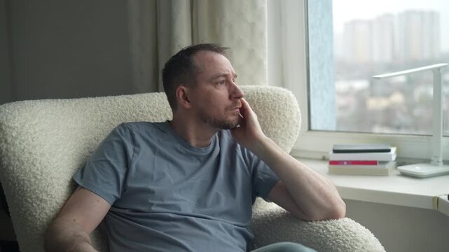 A man sits thoughtfully in a comfortable chair, gazing out the window. He appears contemplative, lost in his own thoughts. The urban skyline is visible in the background.
