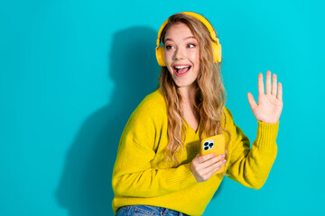 Young woman wearing a bright yellow sweater and headphones waves hello while chatting on a yellow...