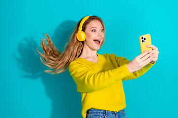 Young woman wearing a bright yellow sweater and headphones takes a joyful selfie against a vivid...
