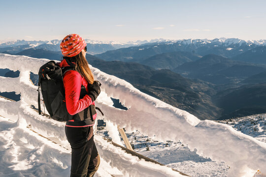 A woman with a backpack gazes at a mountain range from a snow-covered fence. Paganella, Roda refuge,Trentino, Italy
