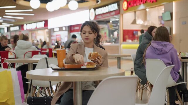 young person dining amidst busy shoppers, student savoring burger in crowded shopping mall setting, casual student relaxing with fast food surrounded by bustling shoppers and urban ambiance