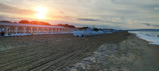 Beach scene at sunset with rows of white sun loungers under white wooden pergola, groomed sand patterns, ocean waves approaching shore, and warm sky colors.