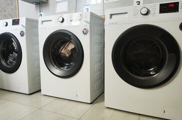 Row of three modern white washing machines with black doors and control panels in laundry room interior. Appliances on tiled floor, ready for use or display.