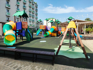 Colorful playground equipment with multiple slides and swing sets on soft rubberized ground surface next to modern multistory building under clear blue sky