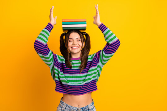Young woman balances colorful books on her head and smiles in yellow studio wear striped sweater playful fashion moment of learning and joy - Powered by Adobe