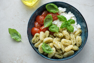 Potato gnocchi with ricotta, roasted tomatoes and fresh basil in a blue bowl, horizontal shot on a light-beige stone background, high angle view