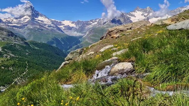 Close-up of crystal clear mountain stream with audio. Fresh green grass and snowy peaks of Matterhorn in background, Zermatt, Switzerland. 4K cinematic nature soundscape. Alpine spring aesthetics.
