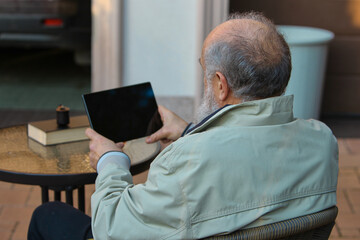 Senior man relaxing outdoors with tablet at patio table in peaceful mood. Concept of lifestyle leisure, senior technology use, home comfort scenes, and calm everyday moments.