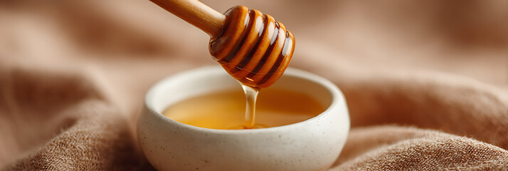 Wooden honey dipper pouring golden liquid honey into a small ceramic bowl against a soft beige fabric background highlighting viscosity and natural sweetness.