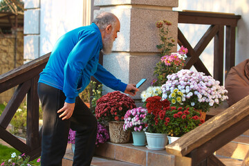 Senior man taking smartphone photo of colorful garden flowers. Concept of documenting beauty, hobby engagement, and digital lifestyle outdoors.