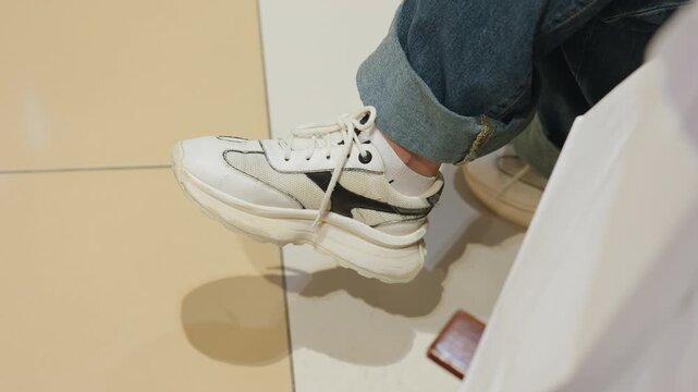 white sneakers on tiled floor, cuffed denim and relaxed posture; candid close-up of shopper sitting by store entrance with phone nearby, subtle shadow and soft mall lighting convey idle waiting