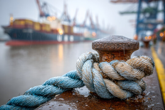 Thick weathered rope tightly secured to a rusty bollard on a wet dock with blurred cargo ships and cranes in the background at a busy industrial harbor during overca