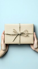 A person's hands holding a wrapped gift tied with twine on a light blue background, top view.