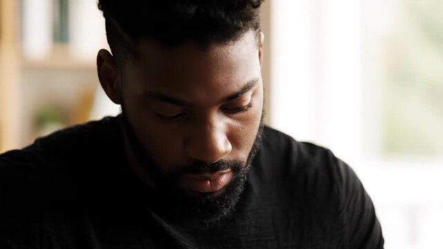 Closeup portrait of a thoughtful young black man with a beard looking down in soft, natural light