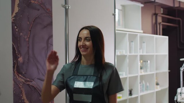 single caucasian stylist standing inside treatment room smiling and waving to incoming client; professional apron, shelves of skincare products, clinical lamp and organized equipment convey modern spa